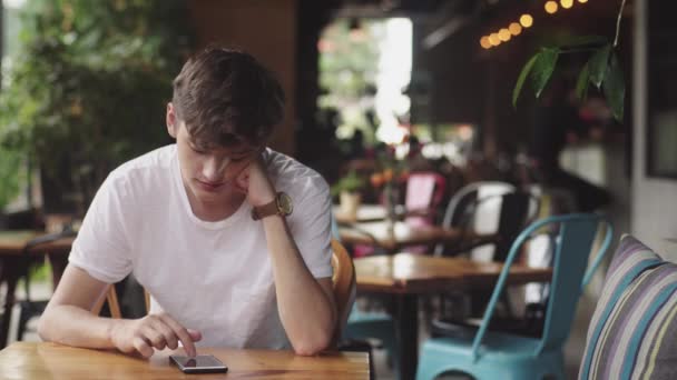 Молодий чоловік серфінгує в Інтернеті в мобільному телефоні, зручно торкається і відправляє текстове повідомлення в кафе. Красивий чат підлітка в мобільному додатку в громадському місці
.  - Кадри, відео
