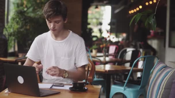Νέος άνθρωπος χρησιμοποιώντας ακουστικά με καλώδια για να απαριθμήσουν τη μουσική στο καφέ, μοντέρνο χώρο εργασίας που κάθονται στο καφέ. Άνοιγμα φορητού υπολογιστή στο εστιατόριο. - Πλάνα, βίντεο