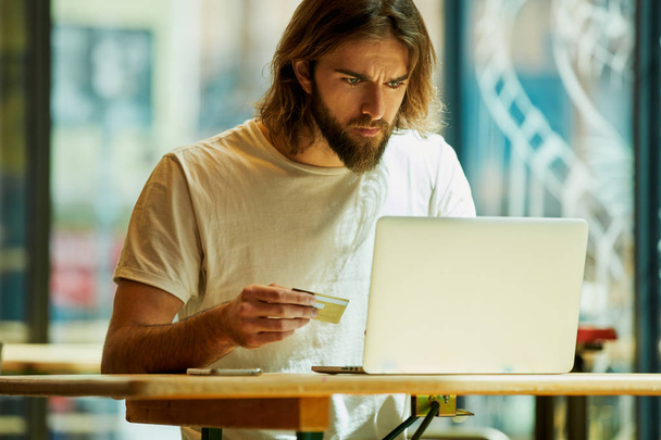 Young bearded handsome man sitting in outside cafe and working with laptop on table - 写真・画像