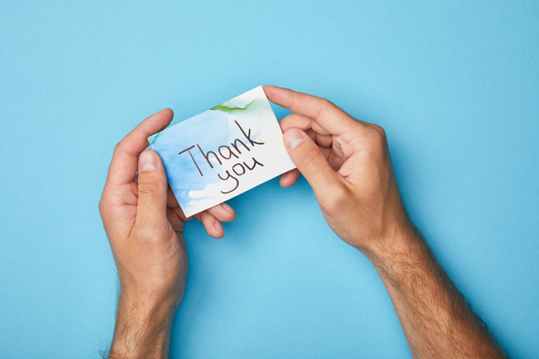 περικομμένη όψη του ανθρώπου κρατώντας ευχετήρια κάρτα με ευχαριστήρια γράμματα σε μπλε φόντο - Φωτογραφία, εικόνα