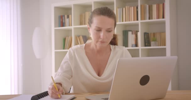 Closeup portrait of elderly caucasian brunette businesswoman using the laptop and taking notes in the office indoors - Кадры, видео