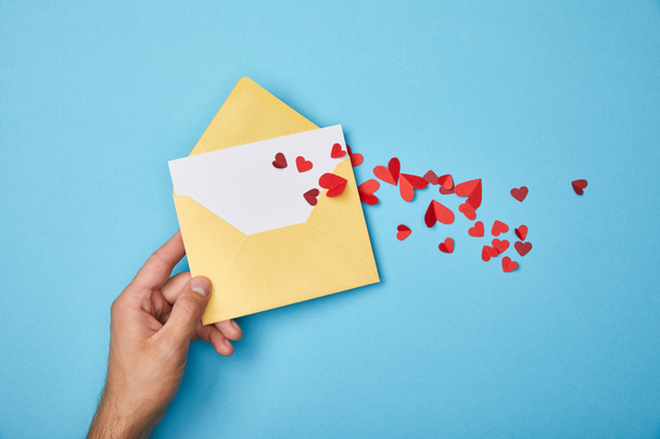 cropped view of man holding yellow envelope with blank white card  and red paper hearts on blue background - Photo, Image