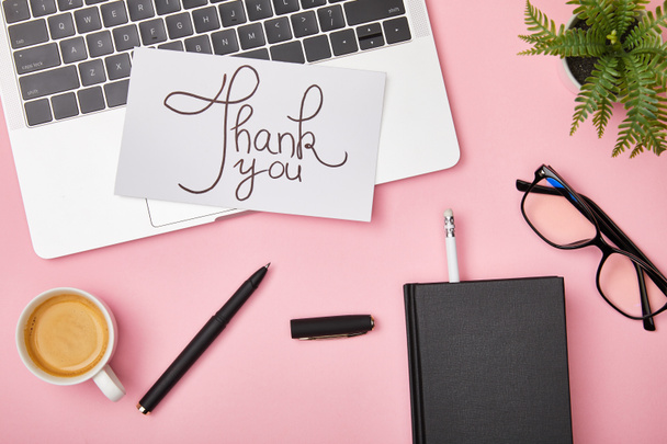 Draufsicht auf Laptop, grüne Pflanze, Kaffee, Notizbuch, Brille und Karte mit Dankesschrift auf rosa Hintergrund - Foto, Bild