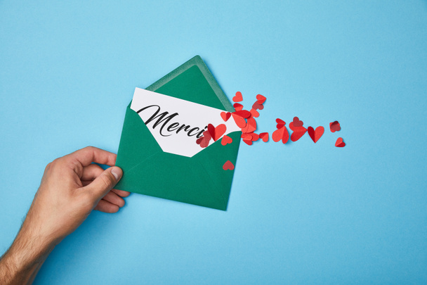 обрезанный вид человека, держащего зеленый конверт с надписью merci на белой карточке и порезанными бумагой красными сердцами на синем фоне
 - Фото, изображение