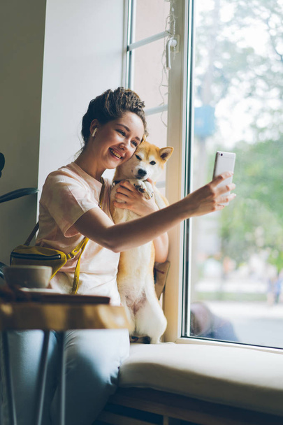 Joyful girl taking selfie in cafe hugging doggy using smartphone on window sill - Фото, зображення
