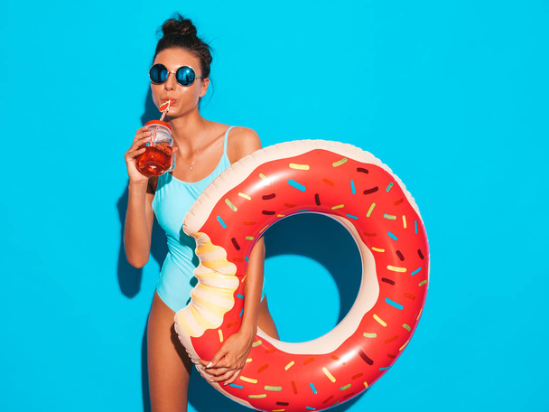 Young beautiful sexy smiling hipster woman in sunglasses.Girl in summer swimwear bathing suit with donut lilo inflatable mattress.Posing near blue wall.Drinking fresh cocktail smoozy drink - Fotoğraf, Görsel