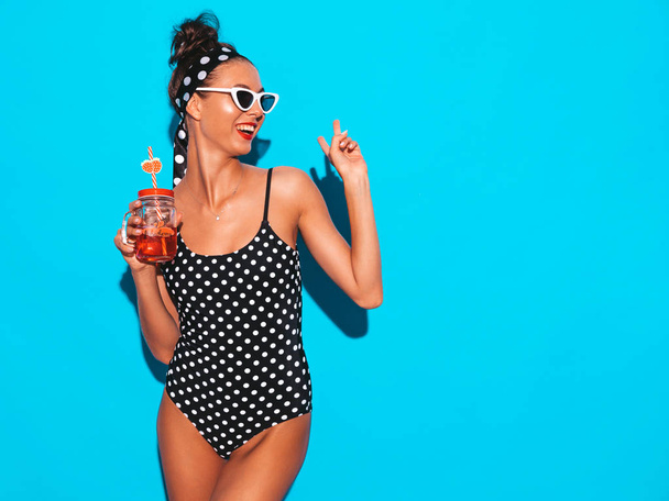Young beautiful sexy smiling hipster woman in sunglasses.Girl in summer peas swimwear bathing suit.Posing near blue wall,drinking fresh cocktail smoozy drink.Shows peace sign - Фото, зображення