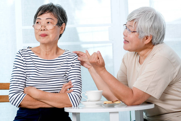Μια ασιατική ηλικιωμένη γυναίκα προσπαθεί να συμφιλιωθεί με την άλλη κατά τη διάρκεια του τσαγιού κοντά στο μπαλκόνι στο σπίτι. - Φωτογραφία, εικόνα
