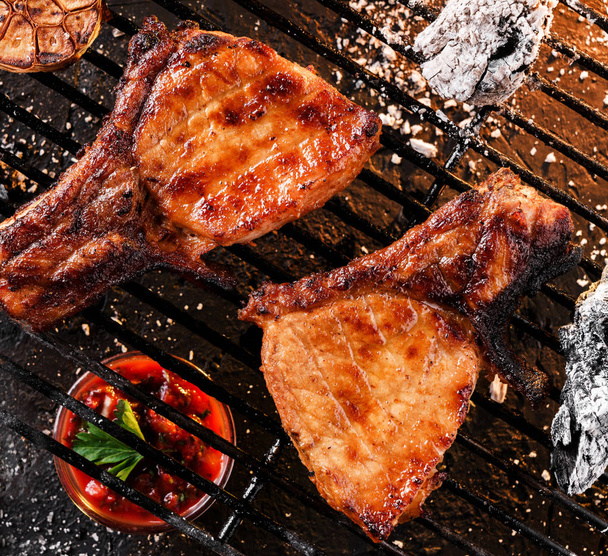 Carne de bife grelhada em T-bone com alho sobre as brasas num churrasco, fundo escuro com luz de fogo. Vista superior
 - Foto, Imagem