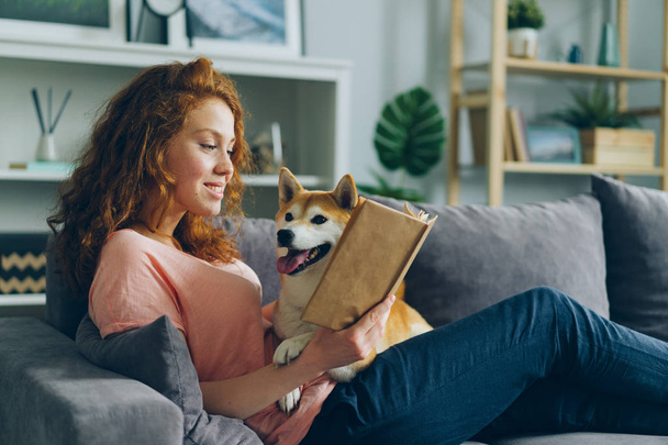 Pretty student reading book in apartment smiling and petting adorable dog - Foto, Imagem