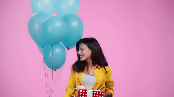 mooi meisje met ballonnen en cadeau kijken naar camera en glimlachend geïsoleerd op roze - Video