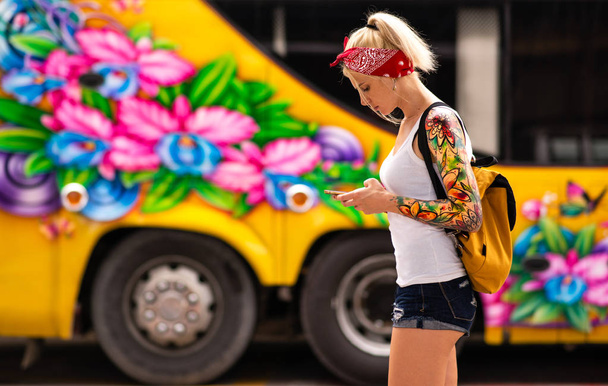 A blonde woman with a phone in hand is standing against the background of a yellow bus. Modern girl wears a red bandage, arms tattooed. - Fotografie, Obrázek