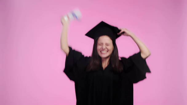 podekscytowany student w akademickiej sukni patrząc na kamerę i skoki z kapelusza ukończenia studiów i dyplom izolowane na różowy - Materiał filmowy, wideo