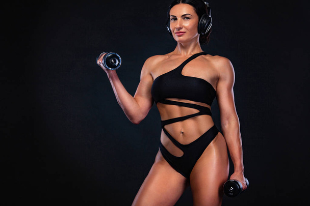 Strong muscular bodybuilder athletic woman pumping up muscles with dumbbells on black background. Workout bodybuilding concept. - Фото, изображение