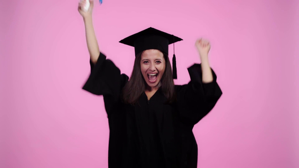 boldog diák tudományos köntös ujjongott összeszokott kézzel és bemutató diploma kamera izolált rózsaszín - Felvétel, videó