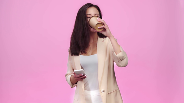 menina bonita usando smartphone, sorrindo e bebendo café para ir isolado em rosa
 - Filmagem, Vídeo