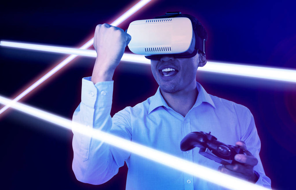 Happy businessman with VR glasses clenching fist while playing video game against gradient - Fotografie, Obrázek