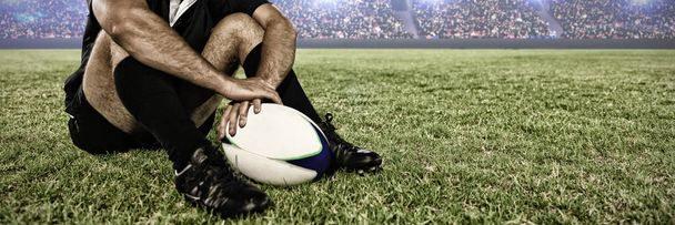 Rugby player sitting on the field with football against digital image of crowded soccer stadium - Photo, Image