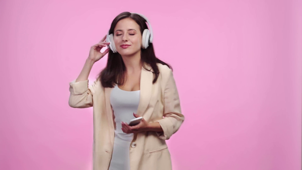 hermosa chica sonriente en auriculares con teléfono inteligente escuchar música y bailar aislado en rosa
 - Metraje, vídeo