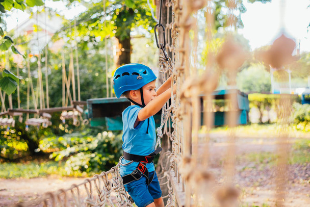 Little cute boy enjoying activity in a climbing adventure park on a summer sunny day.  Climbing extreme sport with helmet and insurance - Zdjęcie, obraz