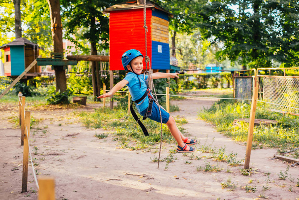 Μικρό χαριτωμένο αγόρι απολαμβάνει δραστηριότητα σε ένα πάρκο αναρρίχησης περιπέτεια σε μια καλοκαιρινή ηλιόλουστη μέρα. Αναρρίχηση ακραίο άθλημα με κράνος και ασφάλεια - Φωτογραφία, εικόνα