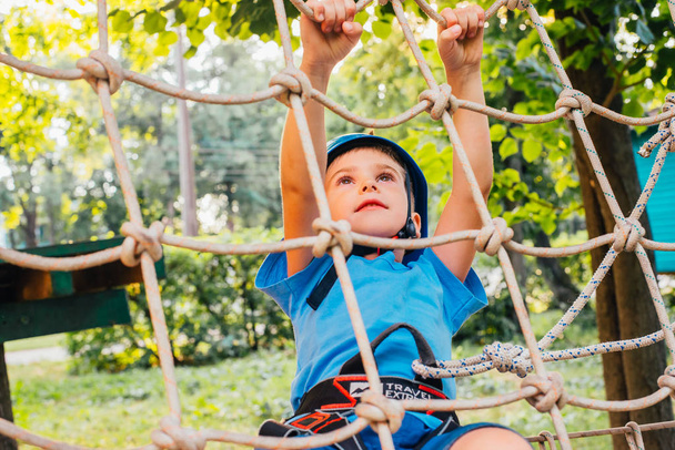 Kleine schattige jongen genieten van activiteit in een klim avonturenpark op een zonnige dag in de zomer. Klimmen extreme sport met helm en verzekeringen - Foto, afbeelding