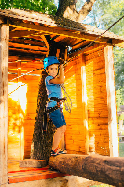 Little cute boy enjoying activity in a climbing adventure park on a summer sunny day.  Climbing extreme sport with helmet and insurance - Foto, imagen