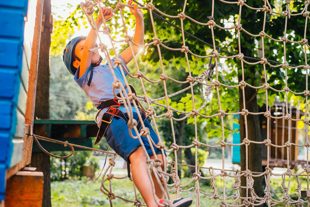 Little cute boy enjoying activity in a climbing adventure park on a summer sunny day.  Climbing extreme sport with helmet and insurance - Fotó, kép