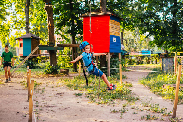 Μικρό χαριτωμένο αγόρι απολαμβάνει δραστηριότητα σε ένα πάρκο αναρρίχησης περιπέτεια σε μια καλοκαιρινή ηλιόλουστη μέρα. Αναρρίχηση ακραίο άθλημα με κράνος και ασφάλεια - Φωτογραφία, εικόνα