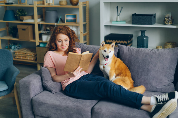Pretty young woman reading book and caressing dog sitting on couch at home - Valokuva, kuva