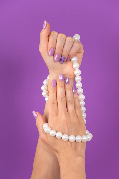 Hermosas manos femeninas con una manicura de color lila con collar de perlas aisladas sobre fondo púrpura en el estudio. Concepto de manicura y belleza. Primer plano, enfoque selectivo
 - Foto, imagen