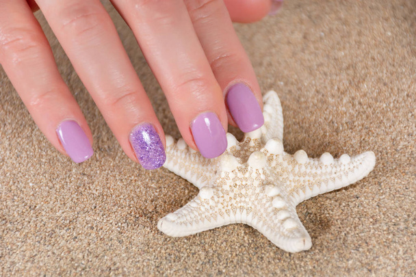 Krásná dívka ruce s fialový barevnou manikúru v pozadí se dotýkejte hvězdného a mořského písku. Koncept manikúry a krásy. Zavřít, selektivní zaměření - Fotografie, Obrázek