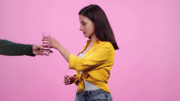 θλιβερό άρρωστο κορίτσι με πονοκέφαλο αγγίζοντας το μέτωπο, παίρνοντας χάπι και το ποτήρι του νερού από τη γυναίκα και κοιτάζοντας την κάμερα απομονωμένη σε ροζ - Πλάνα, βίντεο
