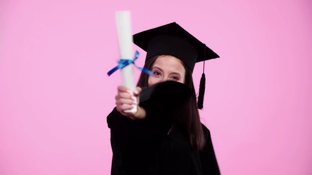 šťastný student v akademických šatech s sevřenými dlaněmi a ukazující diplom na kameru izolovaný on Pink - Záběry, video