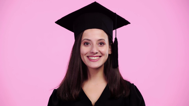 カメラを見て、ピンクに孤立した笑顔のアカデミックガウンの美しい学生 - 映像、動画