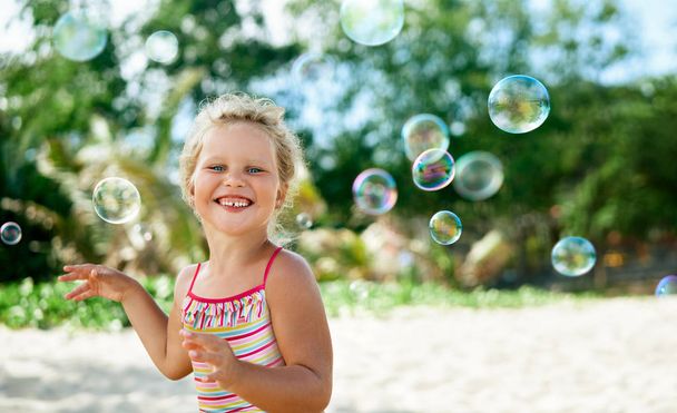 Little happy girl playing soap bubbles on tropical beach. Childhood, lifestyle concept - Photo, Image