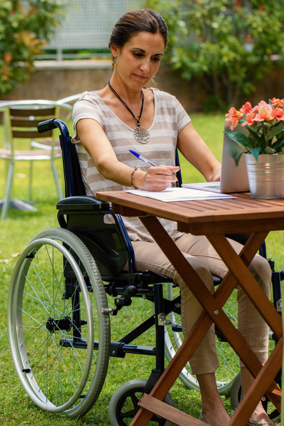 Primer plano retrato de una joven discapacitada en silla de ruedas al aire libre.Mujer sentada a la mesa con portátil, pluma y papel en el jardín
. - Foto, Imagen