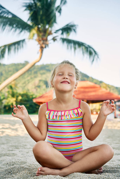 Pretty little girl meditating in Lotus Pose on sandy beach in summer vacation. - Photo, image