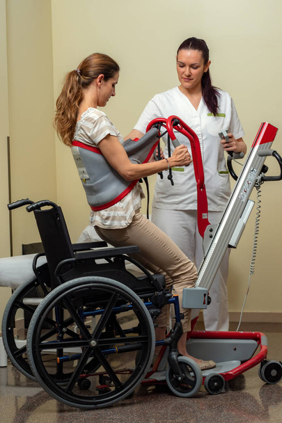 Full length portrait of young female nurse moving handicapped woman with hydraulic lift to wheelchair in hospital. - Foto, imagen