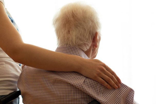 Detalle cercano del brazo femenino abrazando al anciano. Vista trasera de sénior con luz de fondo de ventana
.  - Foto, Imagen