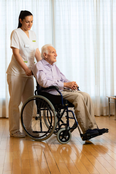 Retrato completo del viejo discapacitado sentado en silla de ruedas. Enfermera joven de pie junto a paciente mayor en la sala de estar
. - Foto, imagen