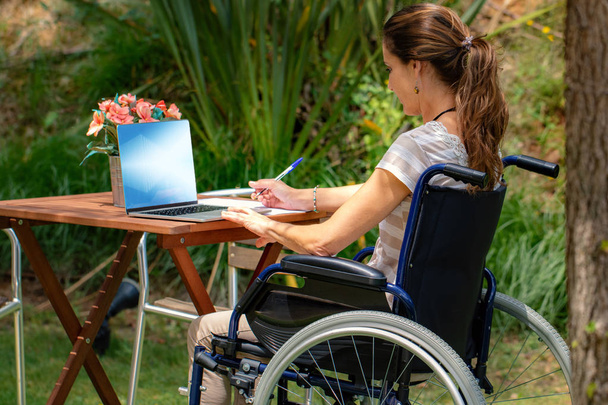 Lähikuva muotokuva nuori halvaantunut nainen pyörätuolissa työskentelevät kannettavan puutarhassa. Takana näkymä tyttö istuu pöydässä ulkona
. - Valokuva, kuva
