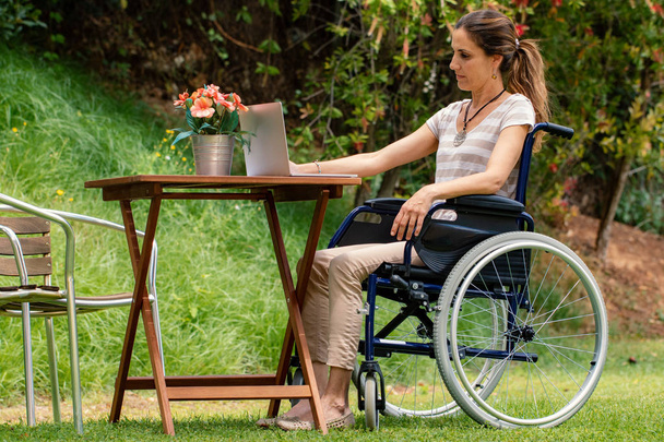 Ολόσωμο πορτρέτο της νεαρής παράλυτης γυναίκας σε αναπηρικό αμαξίδιο που κάθεται στο τραπέζι σε εξωτερικούς χώρους. Πλευρική όψη του κοριτσιού που εργάζεται στο φορητό υπολογιστή στην γκαρντ. - Φωτογραφία, εικόνα