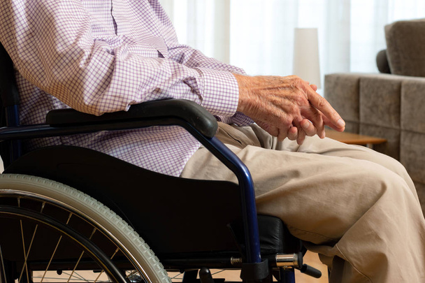Close up conceptual detail shot of old man sitting in wheelchair with hands on lap indoors. - Photo, Image