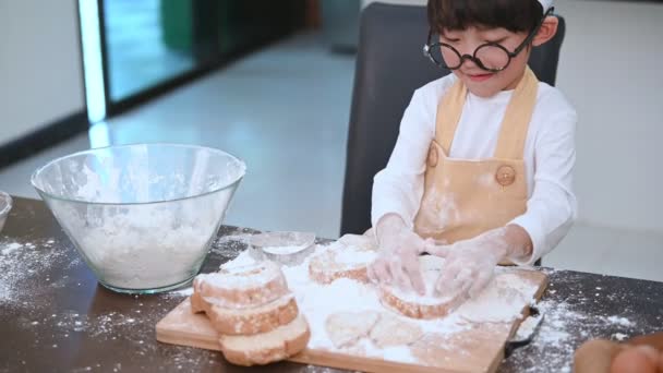 Cute little Asian boy with eyeglasses, chef hat and apron playing and baking bakery in home kitchen funny. Homemade food and bread. Education and learning concept. Portrait and dream job occupation. - Footage, Video