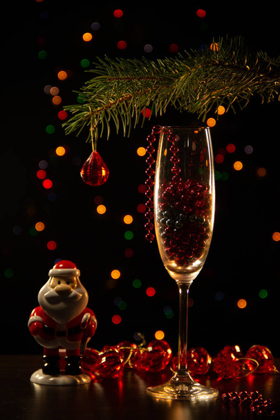 A glass goblet, a new-year decor, a toy Santa Claus, a branch of a Christmas tree on a black background with bright highlights. - Photo, Image