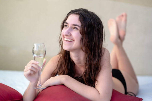 Молодая сексуальная темноволосая женщина в бикини улыбается с бокалом шампанского в гостиной на красной подушке
 - Фото, изображение