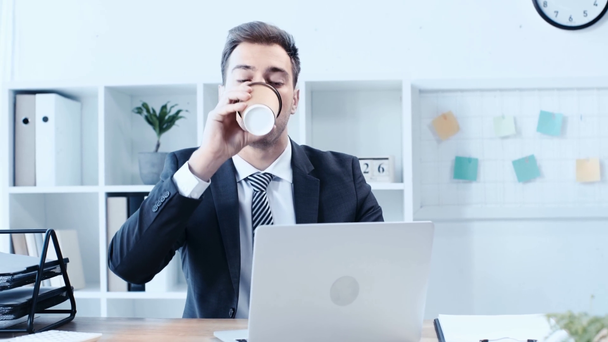 ハンサムな、気配りのビジネスマンは、ラップトップに入力し、職場に座っている間、使い捨てカップからコーヒーを飲みます - 映像、動画