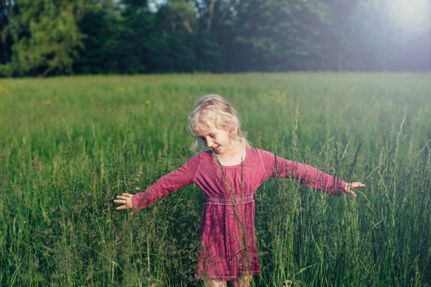 Cute adorable beautiful preschool Caucasian girl walking in tall high grass on meadow at sunset. Happy child kid enjoying summer. Lifestyle authentic childhood. Village rustic rural life. - Photo, Image