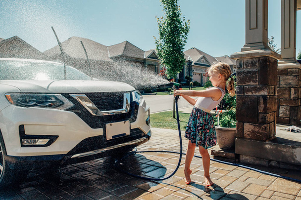 晴れた夏の日にフロントハウスの車道でかわいい幼稚園の小さな白人の女の子が車を洗います。キッズホームの用事義務は責任の概念を家事。ホーススプレー水で遊ぶ子供. - 写真・画像